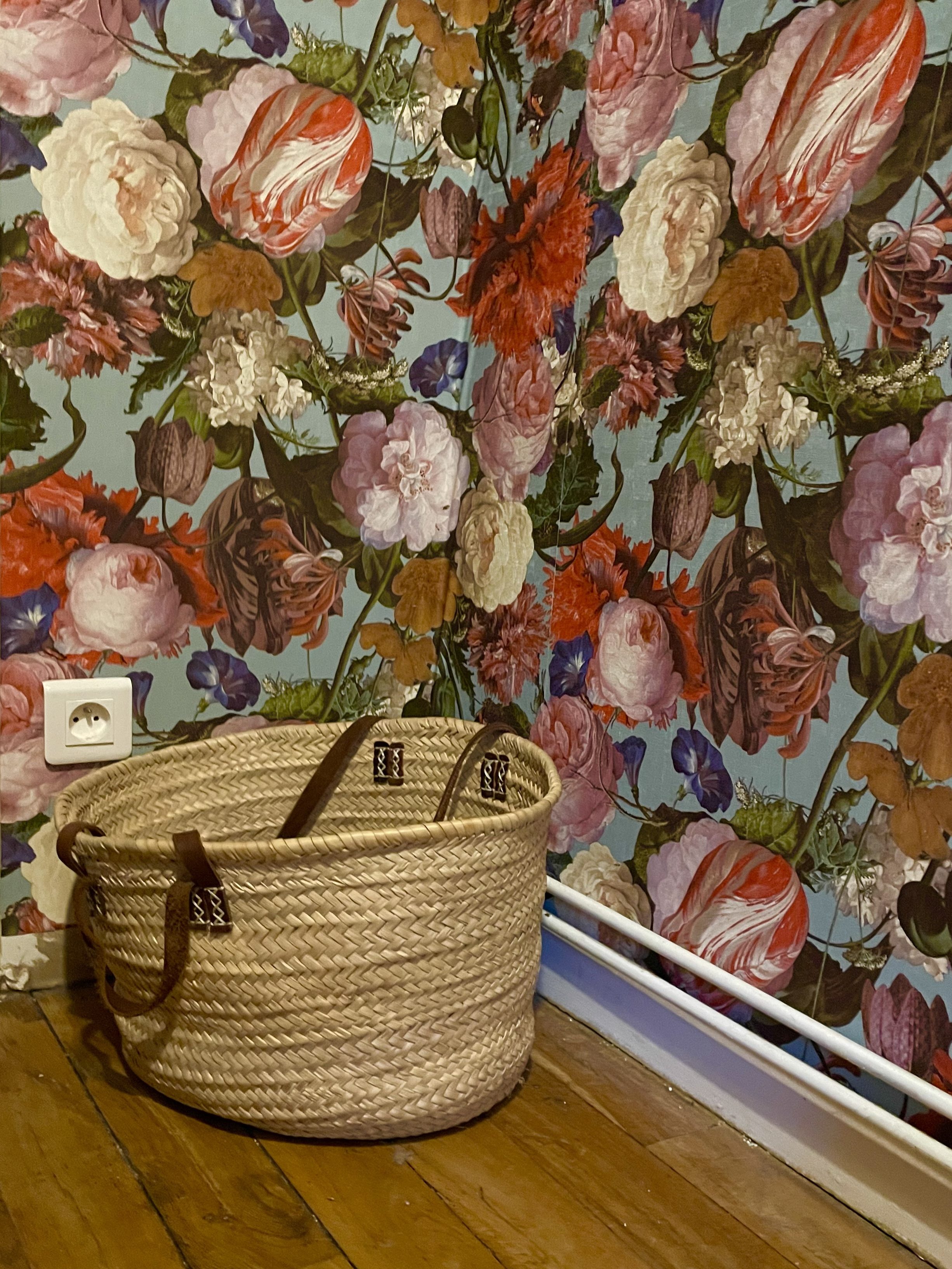Panier à linge vide posé sur parquet devant un papier peint à motif de fleurs